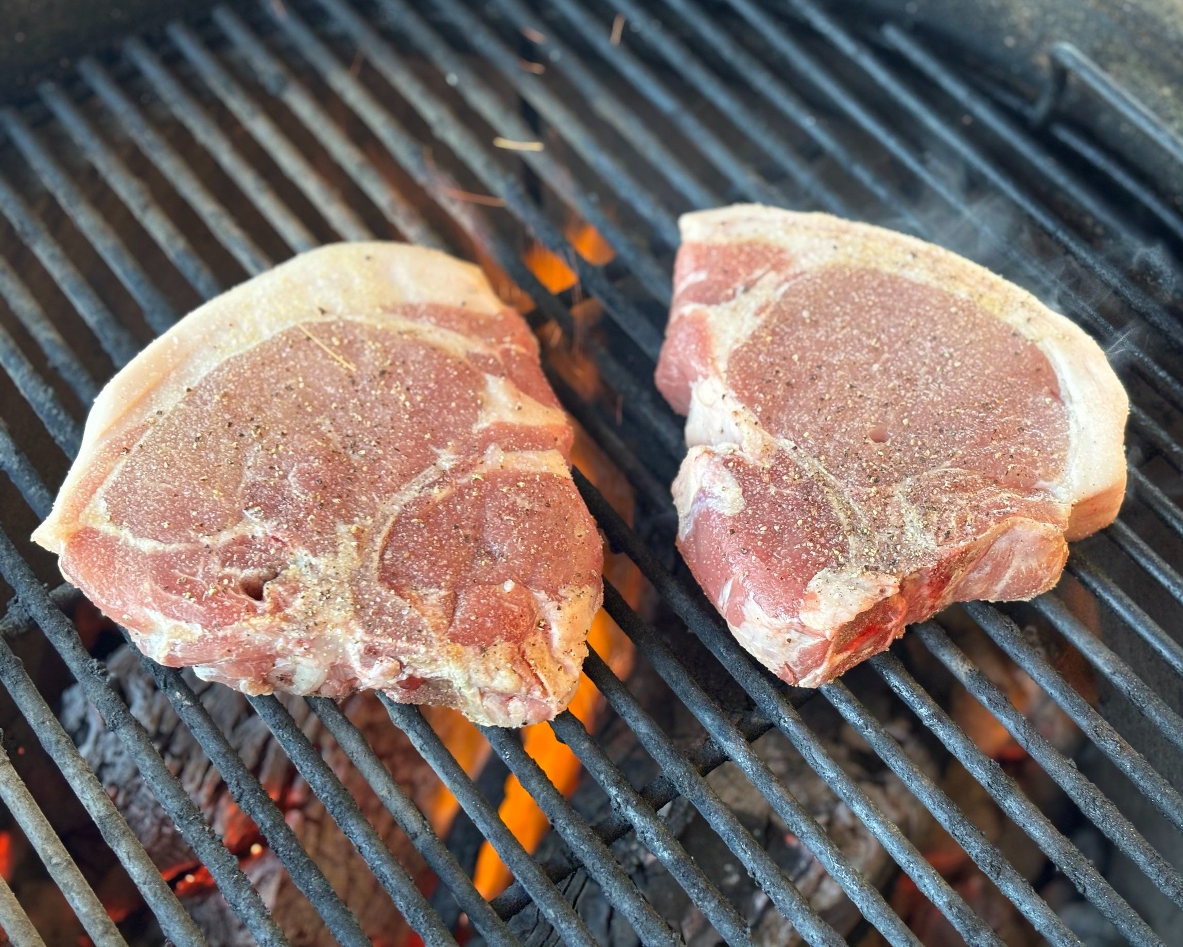 Two seasoned pork chops grilling on a barbecue, with visible grill marks and smoke rising, showcasing a delicious outdoor cooking scene.