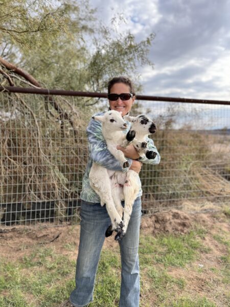 Woman in sunglasses holding two baby goats outdoors, surrounded by greenery and a fence, showcasing farm life and animal care.