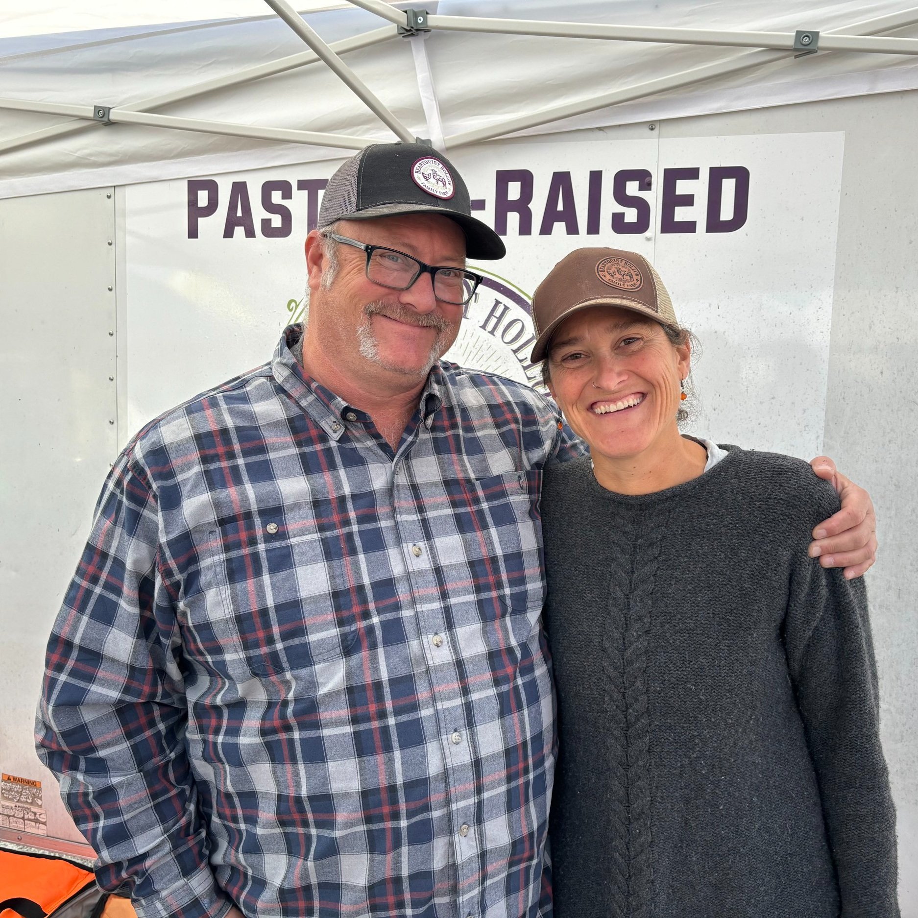 Christie+Heartquist+-+IMG_0798 Smiling man and woman posing together at a farmer's market booth, promoting pasture-raised products, with a white tent and branding visible in the background.