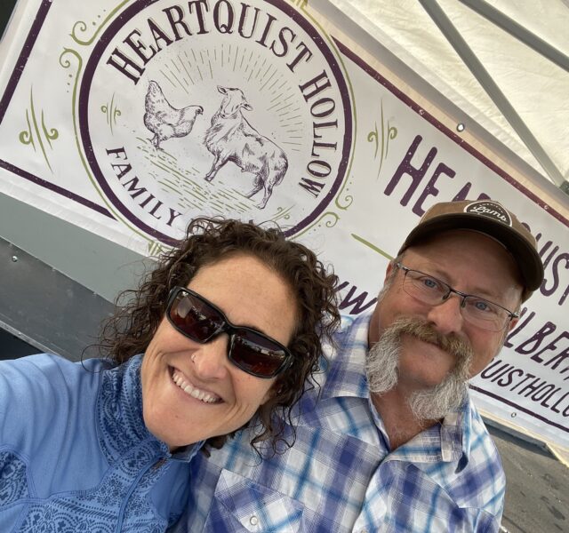 Smiling couple posing in front of the Heartquist Hollow family farm banner, featuring a chicken and a goat illustration, promoting their local agricultural products.