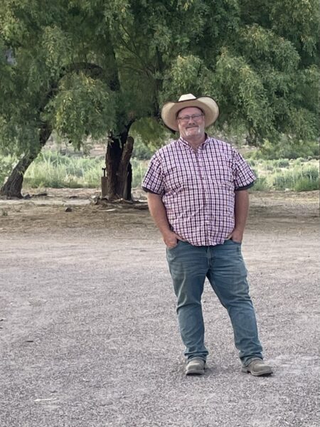 Man in a plaid shirt and cowboy hat standing outdoors, with a tree and rural landscape in the background.