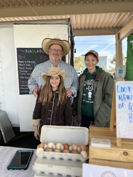 Family at a farmers market stand, featuring a man and two girls wearing cowboy hats. They are smiling in front of a display of fresh eggs and a menu board listing available products like beef and lamb. The setting is sunny and welcoming, showcasing local agriculture and family-run business charm.