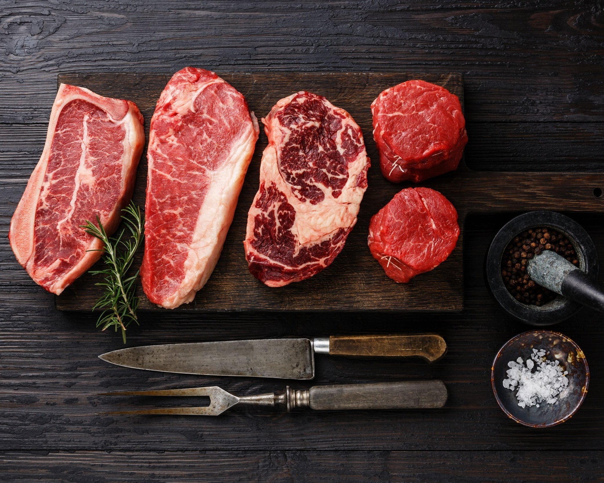 Assorted cuts of raw beef, including ribeye, sirloin, and tenderloin, arranged on a wooden cutting board with fresh rosemary, a mortar and pestle with peppercorns, and bowls of sea salt. Ideal for grilling or gourmet cooking.